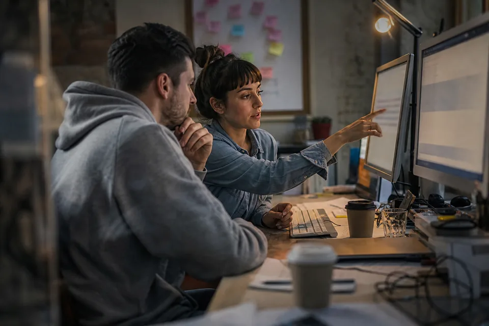Two agency colleagues at a shared desk comparing client chat dashboards on two monitors